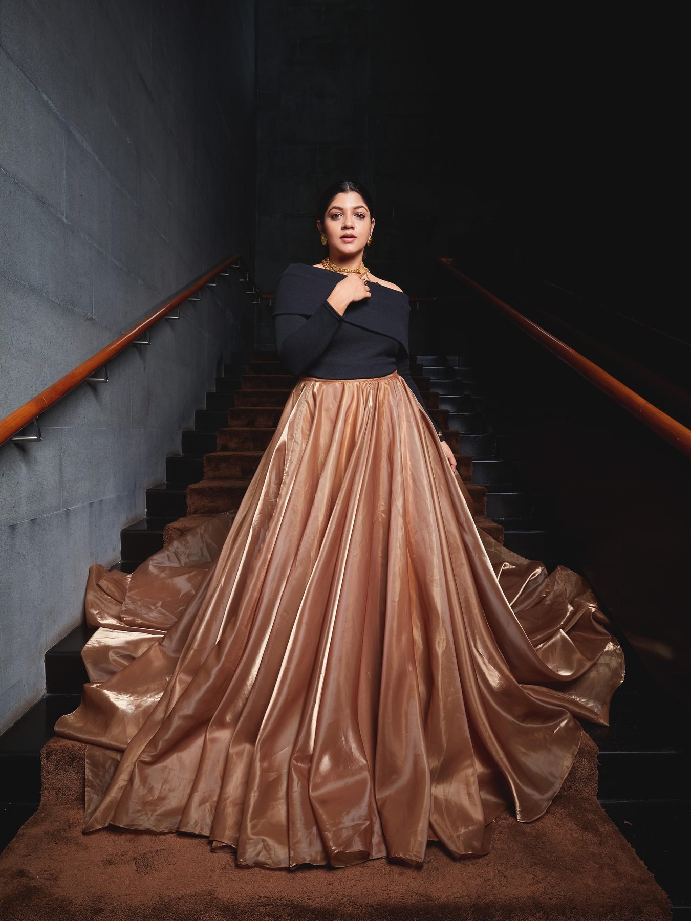 Aparna Balamurali in a black off-shoulder top and gold skirt on a dark staircase, makeup by Anu Rotrico