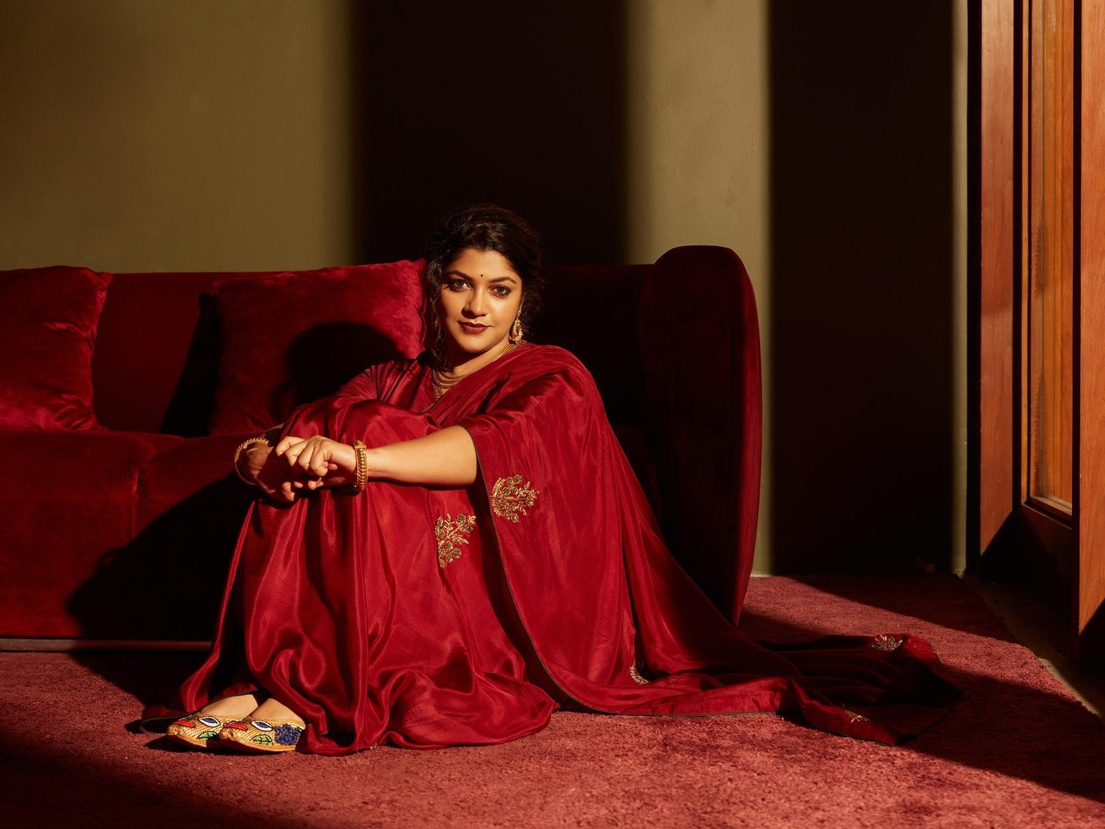 Aparna Balamurali in a deep red silk saree, seated in warm window light on a red velvet sofa