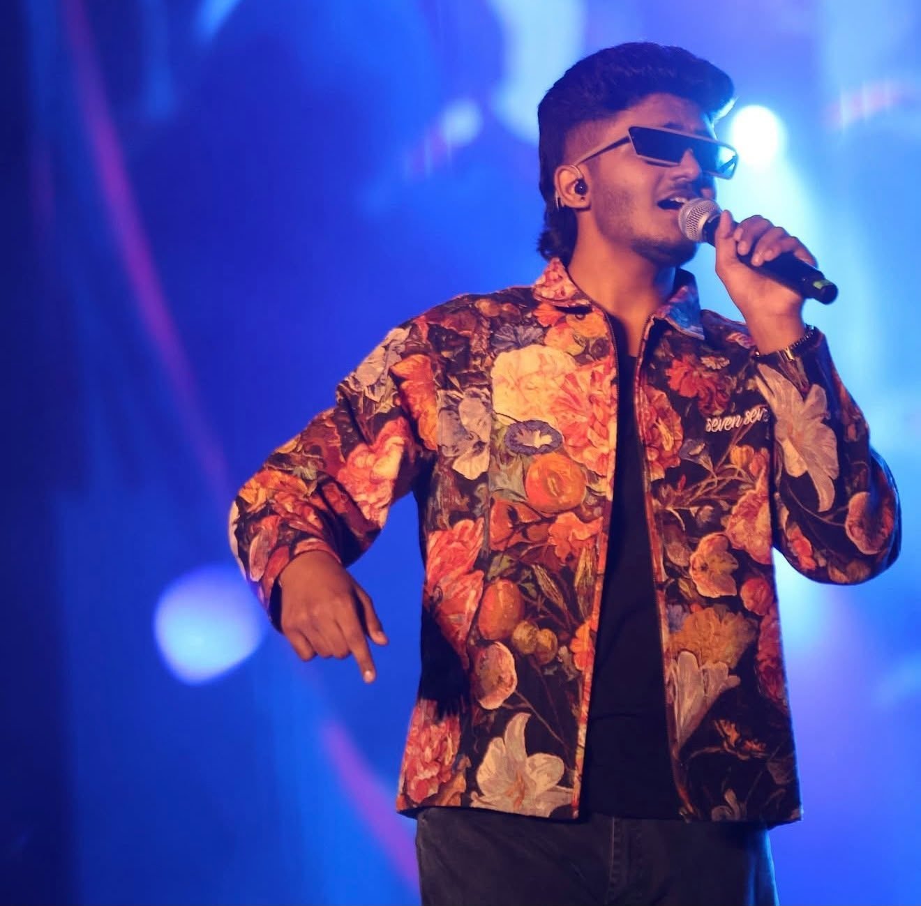 Sai Abhyankkar performing live in a floral tapestry jacket under blue stage light, grooming by Anu Rotrico